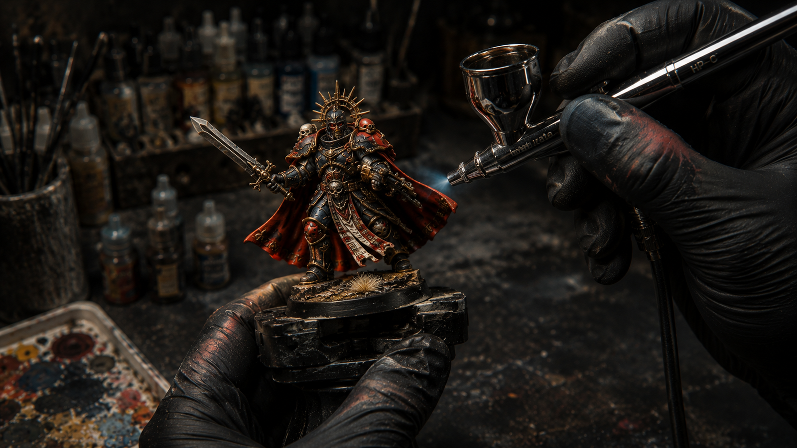Mano pintando una miniatura de Warhammer con aerografo sobre peana negra, con pinturas Citadel y paleta de colores al fondo